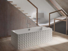 Rechteckige Badewanne aus Stahl-Email mit geometrischer Musterstruktur, freistehend auf Holzfußboden vor Treppe.