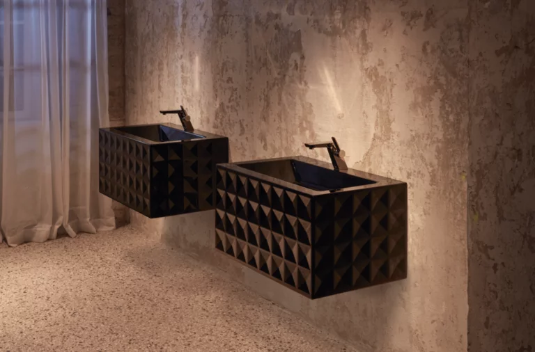 Dos lavabos rectangulares sobre encimeras con frente geométrico junto a pared rugosa, iluminación ambiental.