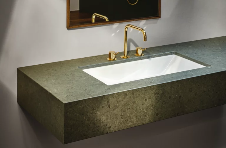 Прямоугольная стальная эмалированная раковина на зелёной столешнице, золотой смеситель, зеркало на заднем плане, современный стиль.