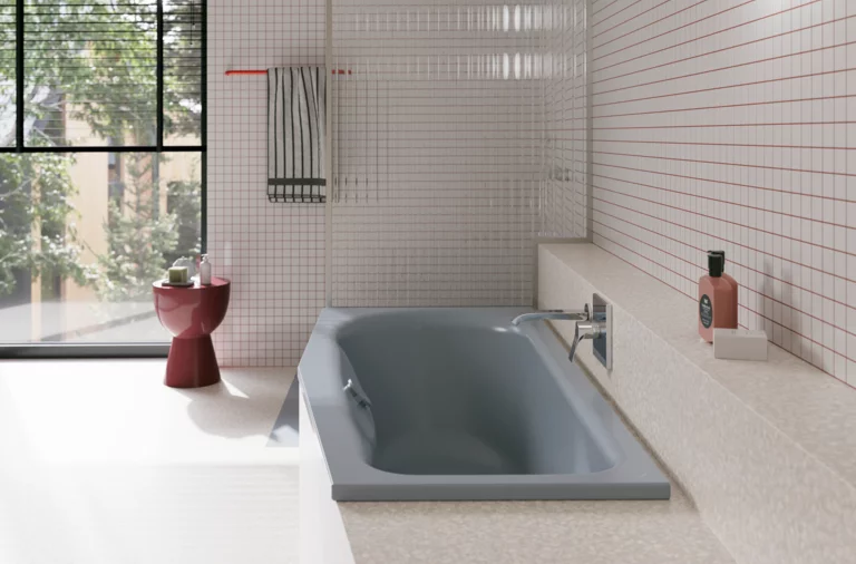 Graue Badewanne in hellem Bad mit weißen Fliesen, großem Fenster, Armatur und rotem Hocker.