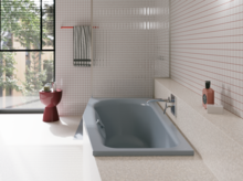 Graue Badewanne in hellem Bad mit weißen Fliesen, großem Fenster, Armatur und rotem Hocker.