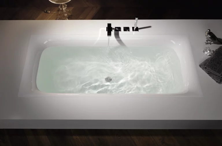 Baignoire rectangulaire en acier émaillé, encastrée dans un panneau blanc, remplie d’eau, robinetterie moderne en haut.