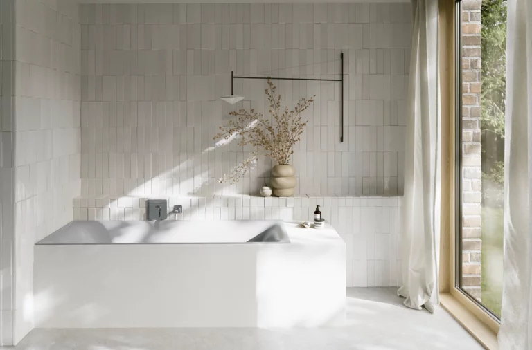 Hellgraue Badewanne in hellem Bad mit weißen Fliesen, Armatur, Vase mit Zweigen und großem Fenster.