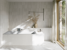 Hellgraue Badewanne in hellem Bad mit weißen Fliesen, Armatur, Vase mit Zweigen und großem Fenster.