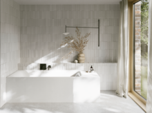 Weiße Badewanne in hellem Bad mit weißen Fliesen, schwarzer Armatur, Vase mit Zweigen und großem Fenster.
