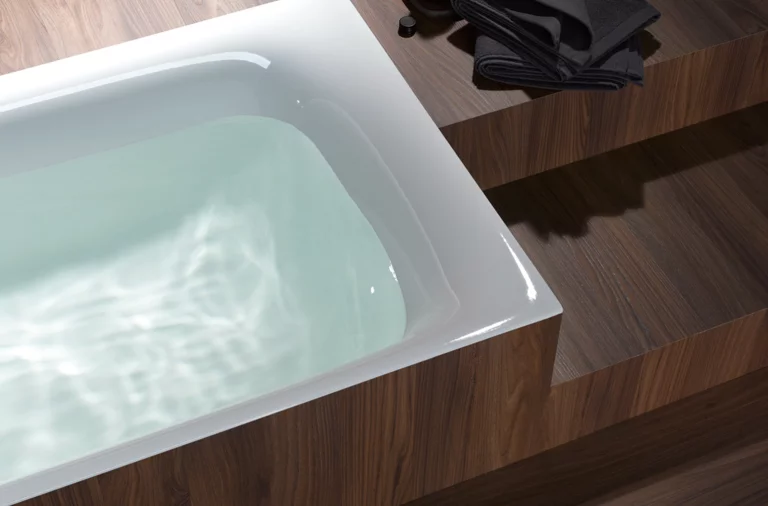 Rechteckige Badewanne aus Stahl-Email, teilweise mit Wasser gefüllt, in Holzverkleidung eingelassen, daneben gefaltete dunkle Handtücher.