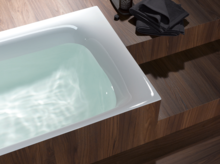 Baignoire rectangulaire en acier émaillé partiellement remplie d’eau, encastrée dans un habillage bois, serviettes foncées pliées à côté.