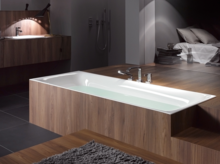 Rechteckige Stahl-Email-Badewanne, halb eingelassen in Holzverkleidung, daneben Waschbecken auf Einbauplatte und moderne Badezimmerausstattung.