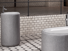 Runde Badewanne und rundes Waschbecken aus Stahl-Email, beide mit grauer Umhüllung, auf gemustertem Fliesenboden.