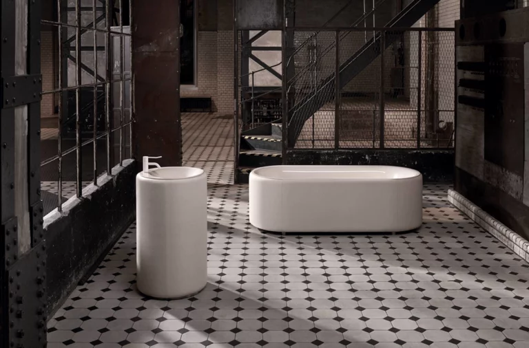 Ovale Badewanne aus Stahl-Email und rundes Standwaschbecken auf gemustertem Fliesenboden in industriellem Ambiente.