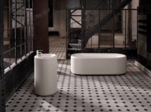 Ovale Badewanne aus Stahl-Email und rundes Standwaschbecken auf gemustertem Fliesenboden in industriellem Ambiente.