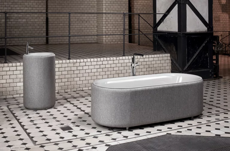 Vrijstaand ovaal bad van staal-emaille en rond vrijstaand wastafel, beide met grijze bekleding op tegelvloer.