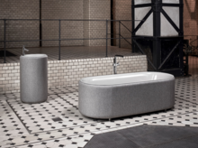 Vrijstaand ovaal bad van staal-emaille en rond vrijstaand wastafel, beide met grijze bekleding op tegelvloer.