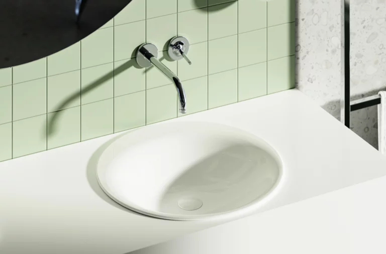 Oval white inset washbasin in a white surface, in front of green tiled wall with round wall-mounted faucet.