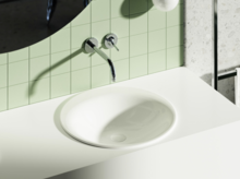 Oval white inset washbasin in a white surface, in front of green tiled wall with round wall-mounted faucet.