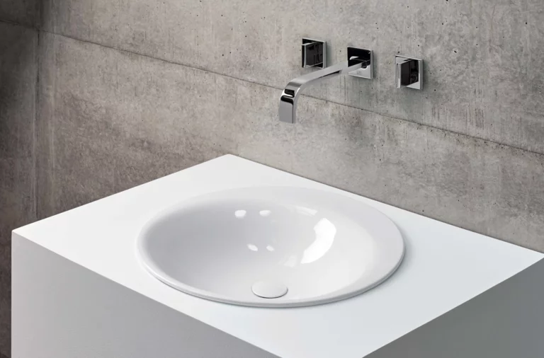 Ronde wastafel van staal-email op wit inbouwblad, wandkraan voor grijze betonnen wand.