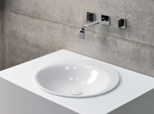 Ronde wastafel van staal-email op wit inbouwblad, wandkraan voor grijze betonnen wand.