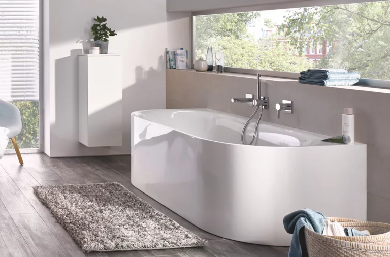 Bañera exenta asimétrica de acero vitrificado en baño moderno con gran ventana y suelo efecto madera.