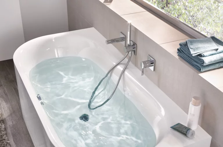 Bañera rectangular de acero esmaltado con agua, desagüe central, grifería moderna y ducha de mano, sobre suelo de baldosas claras.