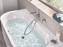 Bañera rectangular de acero esmaltado con agua, desagüe central, grifería moderna y ducha de mano, sobre suelo de baldosas claras.