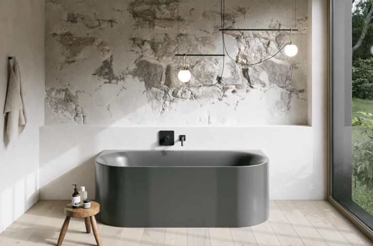 Graue Badewanne in hellem Bad mit Steinoptik-Wand, schwarzer Armatur, Pendelleuchten und großem Fenster.