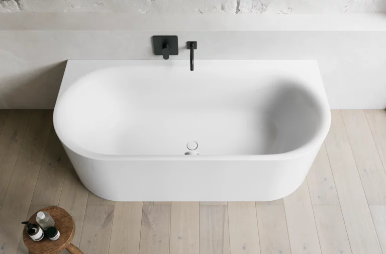 Weiße freistehende Badewanne vor heller Wand mit schwarzer Armatur und Holzfußboden.