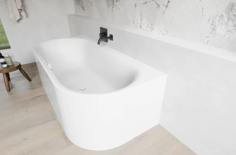 Weiße freistehende Badewanne vor heller Steinoptik-Wand mit schwarzer Armatur und Holzfußboden.