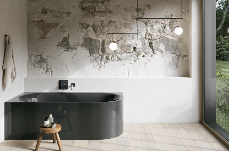 Dunkelgraue Badewanne in hellem Bad mit Steinoptik-Wand, schwarzer Armatur, Pendelleuchten und großem Fenster.