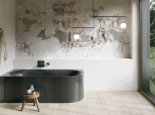 Dunkelgraue Badewanne in hellem Bad mit Steinoptik-Wand, schwarzer Armatur, Pendelleuchten und großem Fenster.