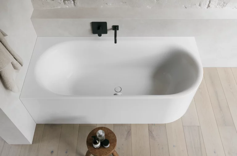 Weiße freistehende Badewanne vor heller Wand mit schwarzer Armatur, Holzfußboden und kleinem Hocker.