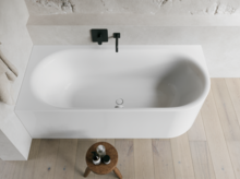Weiße freistehende Badewanne vor heller Wand mit schwarzer Armatur, Holzfußboden und kleinem Hocker.