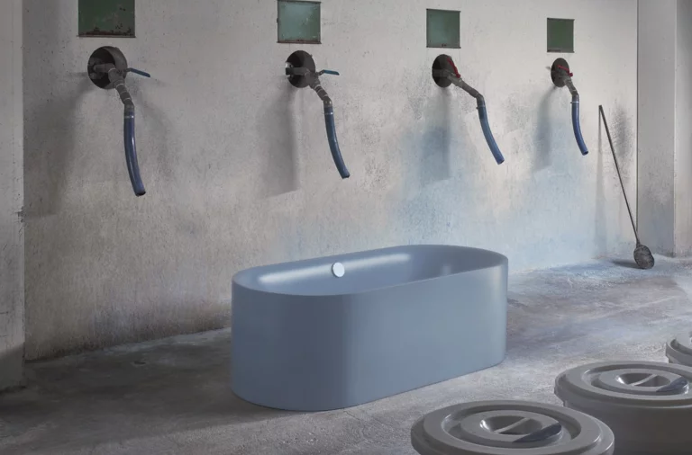 Freistehende, ovale Badewanne aus Stahl-Email in einem industriellen Raum, Wasserhähne an der Wand darüber.
