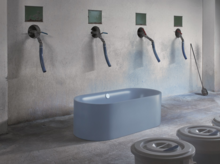 Freistehende, ovale Badewanne aus Stahl-Email in einem industriellen Raum, Wasserhähne an der Wand darüber.