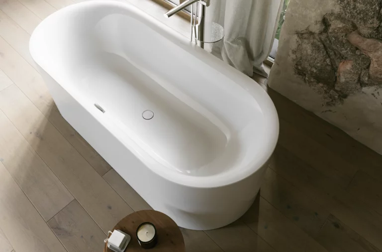 Weiße freistehende Badewanne mit silberner Armatur, hellem Holzfußboden und Steinoptik-Wand.