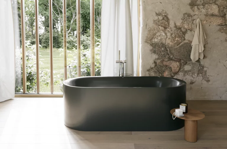 Dunkelgrüne freistehende Badewanne in hellem Bad mit großem Fenster, Steinoptik-Wand und Hocker.