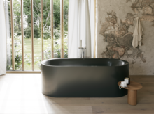 Dunkelgrüne freistehende Badewanne in hellem Bad mit großem Fenster, Steinoptik-Wand und Hocker.