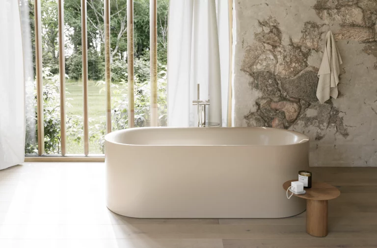 Cremefarbene freistehende Badewanne in hellem Bad mit großem Fenster, Steinoptik-Wand und Hocker.