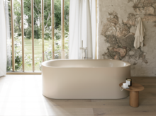 Cremefarbene freistehende Badewanne in hellem Bad mit großem Fenster, Steinoptik-Wand und Hocker.