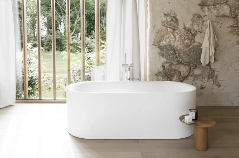 Weiße freistehende Badewanne in hellem Bad mit großem Fenster, Steinoptik-Wand und Hocker.