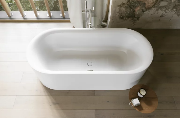 Weiße freistehende Badewanne von oben mit hellem Holzfußboden, großem Fenster und rundem Hocker.