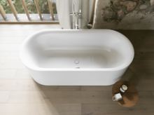 Weiße freistehende Badewanne von oben mit hellem Holzfußboden, großem Fenster und rundem Hocker.