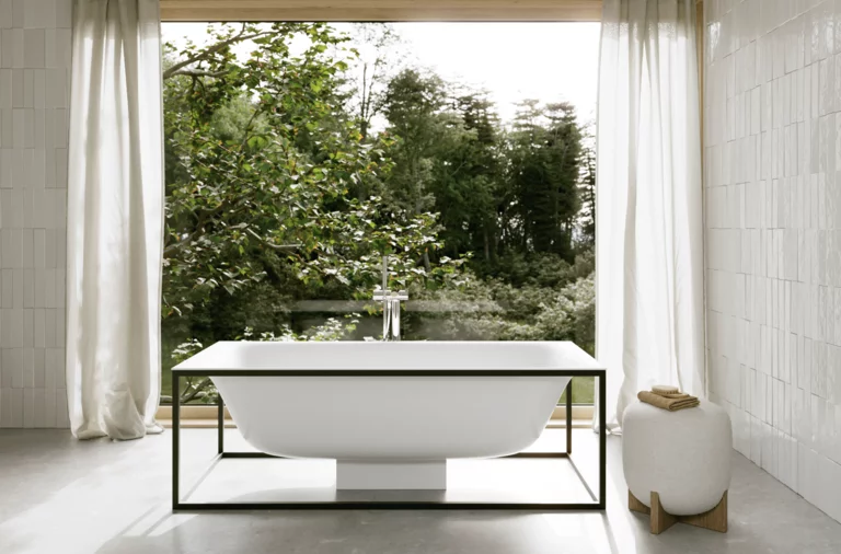 Freistehende weiße Badewanne vor großem Fenster mit Blick ins Grüne, helle Vorhänge und weiß geflieste Wände.