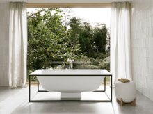 Freistehende weiße Badewanne vor großem Fenster mit Blick ins Grüne, helle Vorhänge und weiß geflieste Wände.