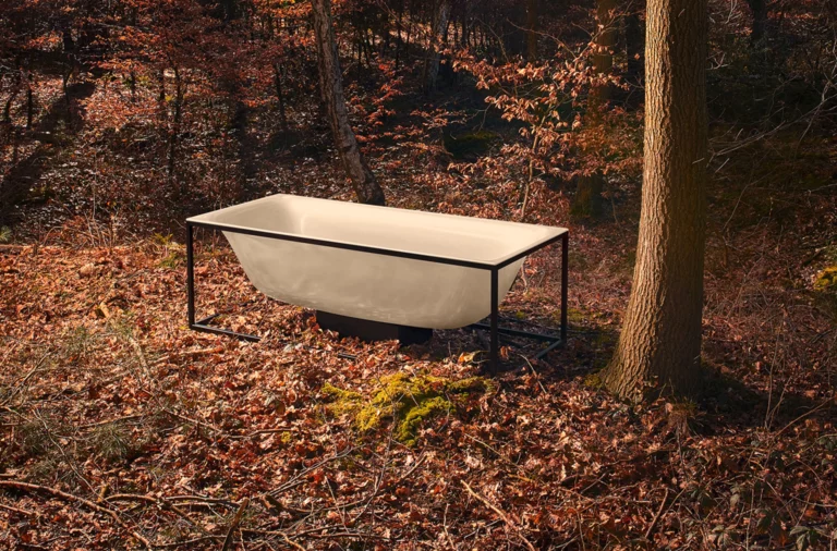 Rectangular steel-enamel bathtub with black frame stands in autumn forest next to a tree.