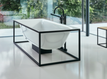 Отдельностоящая прямоугольная ванна из стальной эмали в черном металлическом каркасе, рядом табурет, на фоне большие окна.