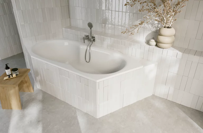 Weiße Einbau-Badewanne in hell gefliester Ecke mit Armatur, Ablagefläche und dekorativer Vase mit Zweigen.