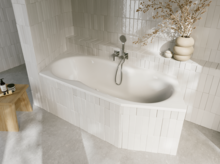 Weiße Einbau-Badewanne in hell gefliester Ecke, mit Armatur, Holzablage und dekorativer Vase mit Zweigen.