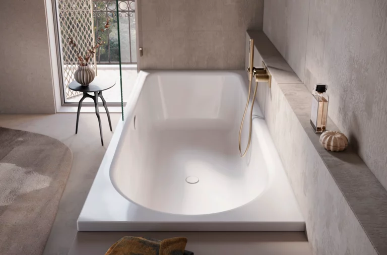 Große Dusch-Badewanne mit goldener Armatur, Ablage, Handtuch und Blick durch das Fenster auf Balkon mit Tisch und Vase.