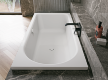 Weiße Badewanne in grauer Badezimmernische mit schwarzer Armatur und heller Holzoptik im Raum.