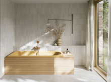 Beigefarbene Badewanne mit gerillter Front in hellem Bad mit weißen Fliesen und großem Fenster.